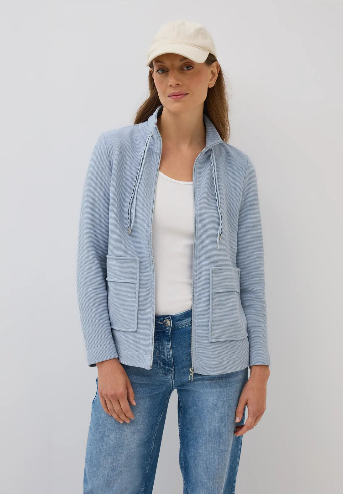 Woman wearing a light blue jacket, white shirt, and jeans on a plain background