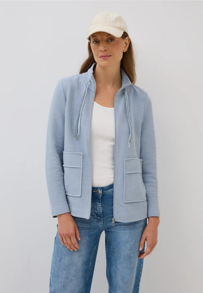 Woman wearing a light blue jacket, white shirt, and jeans on a plain background