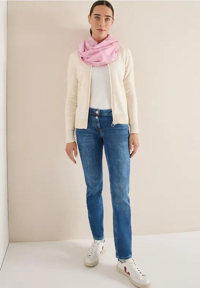 Woman wearing a white cardigan, pink scarf, white shirt, blue jeans, and white sneakers against a beige wall.