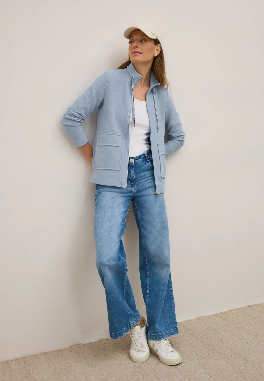 Woman wearing a light blue jacket, white shirt, blue jeans, and white sneakers against a plain background