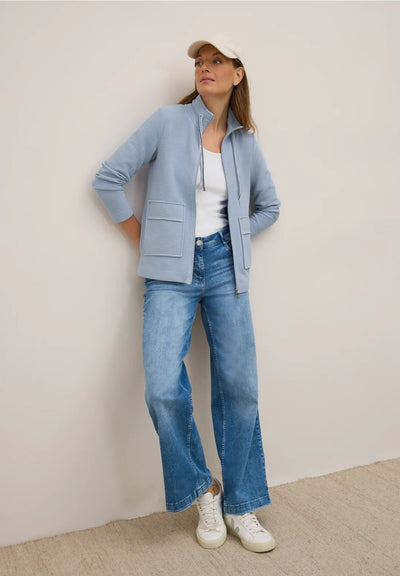 Woman wearing a light blue jacket, white shirt, blue jeans, and white sneakers against a plain background