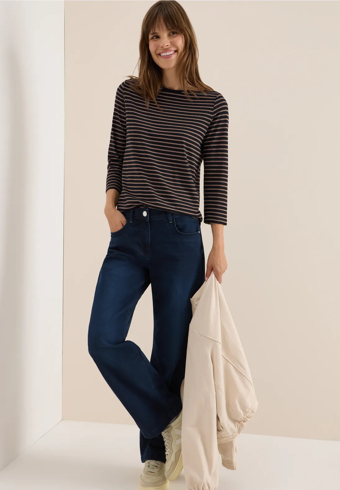 Woman wearing a striped shirt and blue jeans, holding a beige jacket against a neutral background