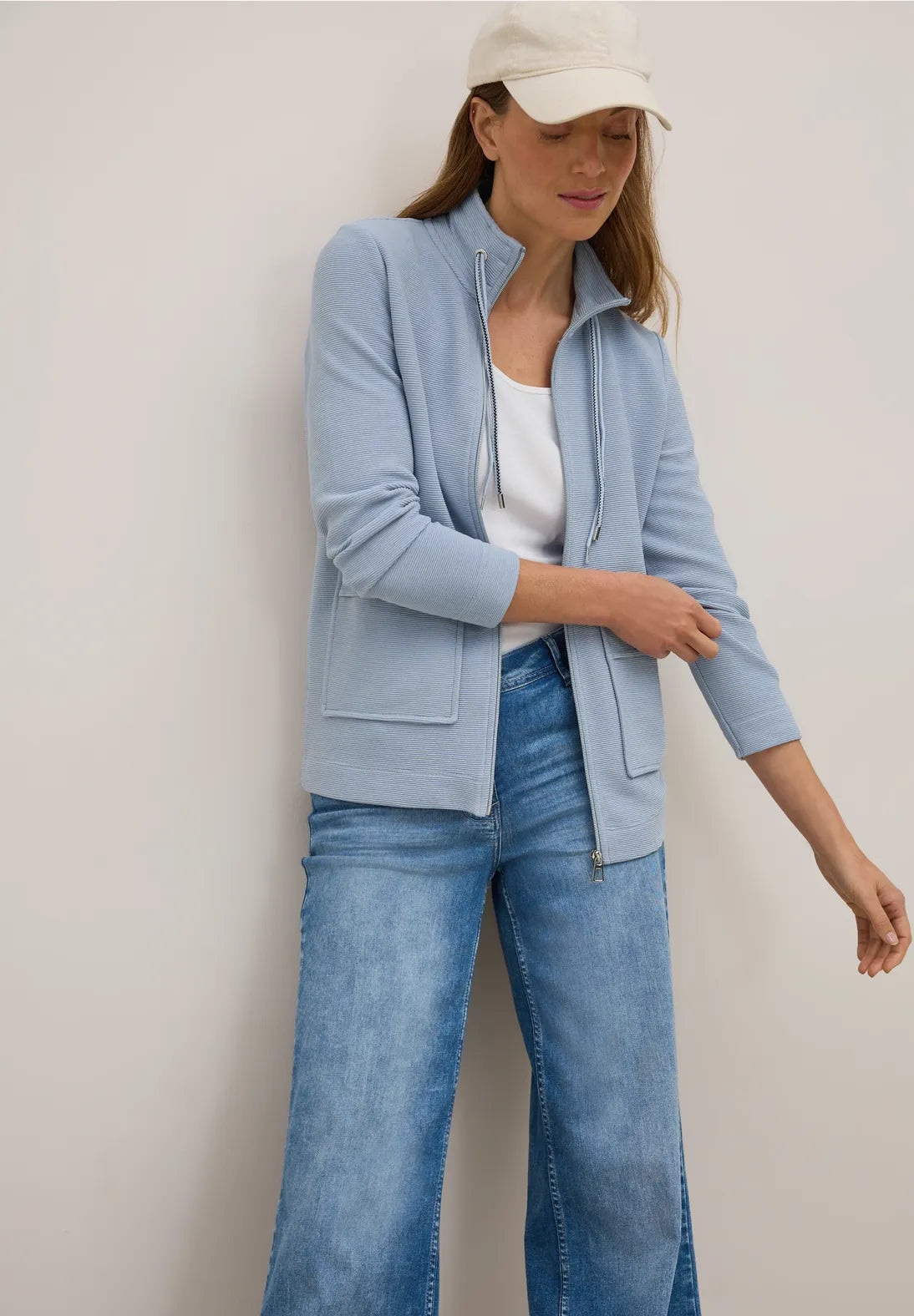 Woman wearing a light blue jacket, white top, and jeans against a plain background