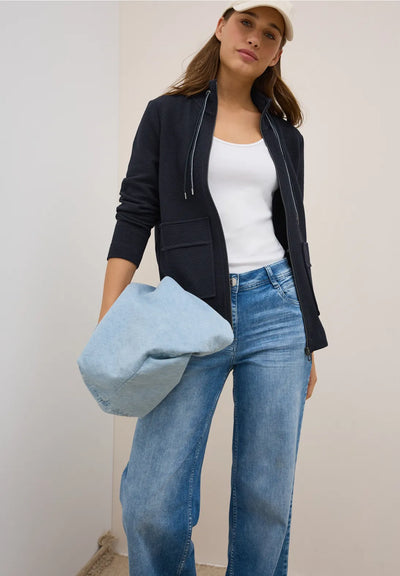 Woman wearing a black jacket, white top, and blue jeans, holding a light blue bag against a plain background.