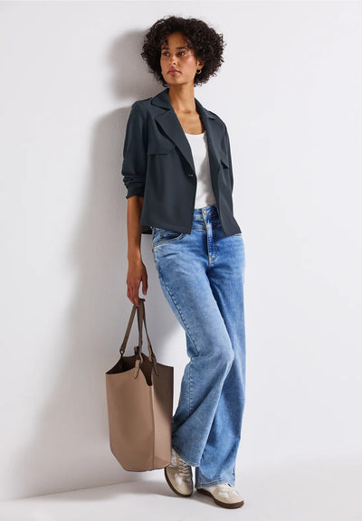 Woman wearing a dark jacket, white shirt, blue jeans, and holding a beige tote bag against a light background