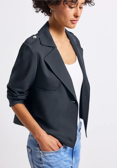 Woman wearing a black blazer over a white top with blue jeans on a light gray background