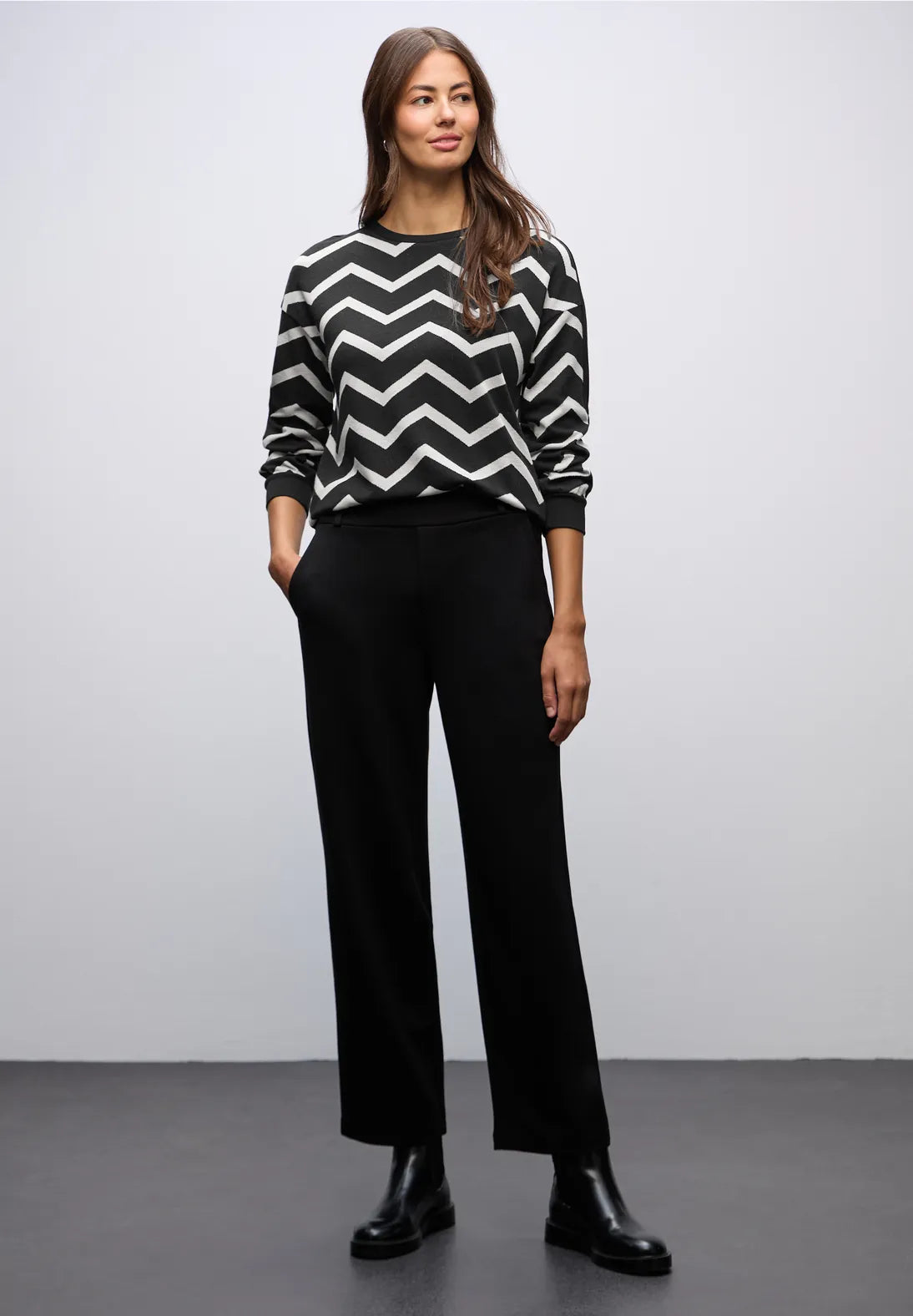 Woman wearing a black and white zigzag sweater with black pants against a plain background