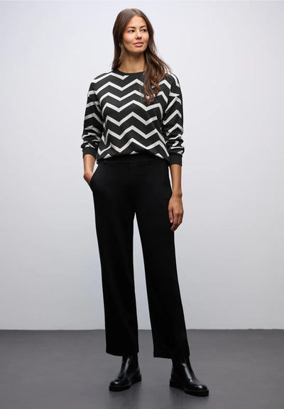 Woman wearing a black and white zigzag sweater with black pants against a plain background