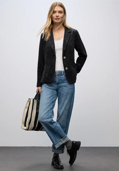 Woman wearing a black blazer, white top, blue jeans, and holding a striped bag against a plain background