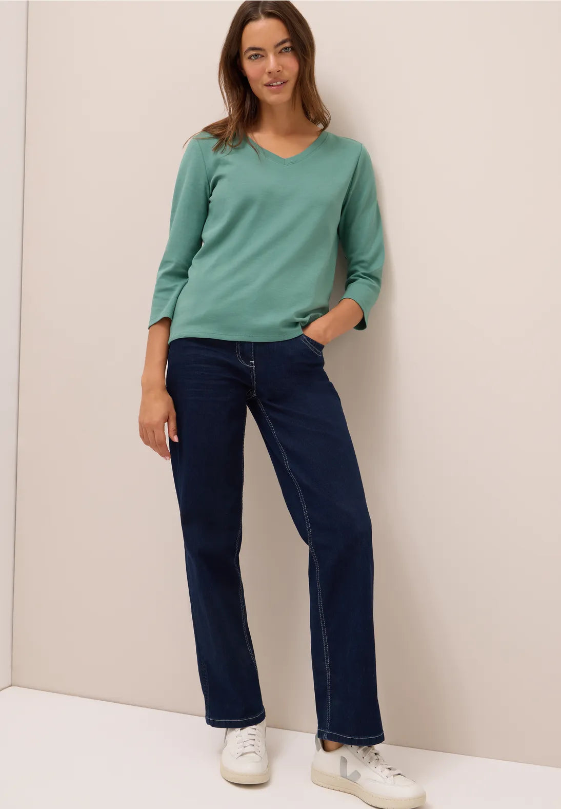 Woman wearing a green long-sleeve top and blue jeans against a plain background