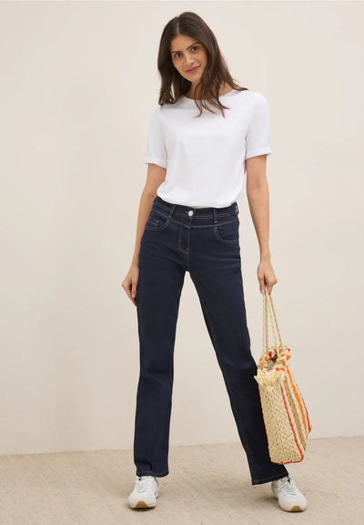 Woman wearing a white t-shirt and dark jeans, holding a straw bag against a plain background
