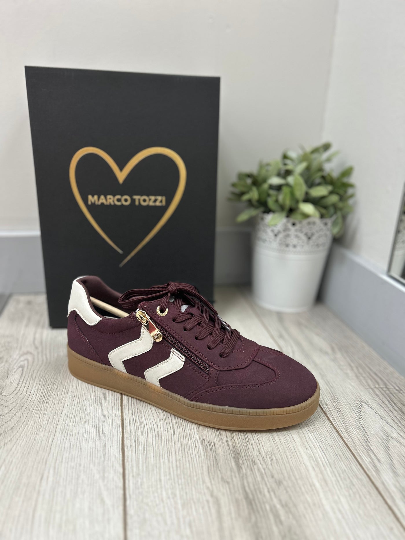 Maroon sneaker with white accents on a light wooden floor, next to a black box with 'Marco Tozzi' branding.
