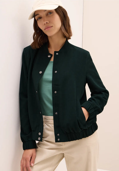 Woman wearing a dark green varsity jacket over a teal shirt and beige pants, standing against a white wall.