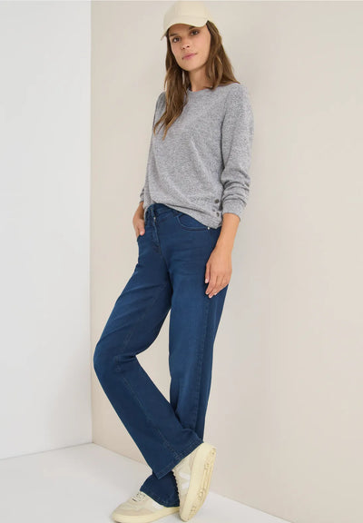 Woman wearing a gray sweater and blue pants against a white wall