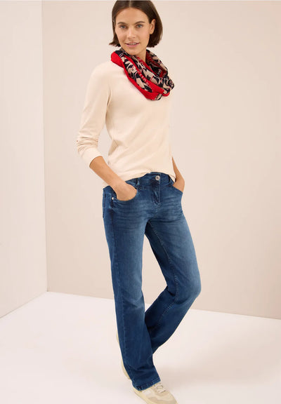Woman wearing a cream sweater and blue jeans with a patterned scarf on a plain background