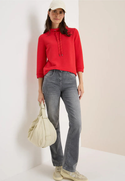 Woman wearing a red sweater, gray jeans, and a white cap, holding a beige bag against a plain background.