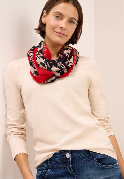 Woman wearing a cream sweater and blue jeans with a colorful patterned scarf around her neck against a plain background