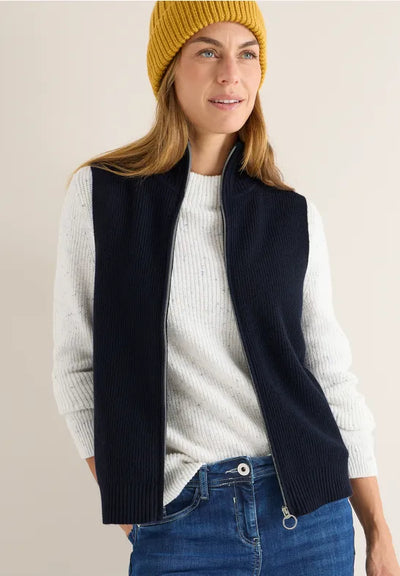 Woman wearing a navy vest over a white sweater with blue jeans and a yellow beanie on a beige background