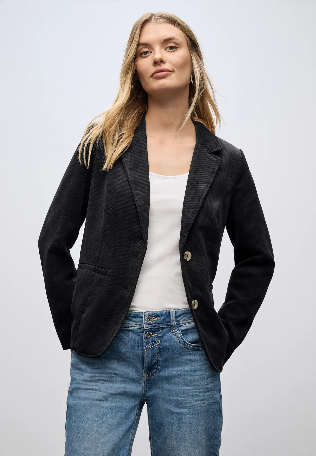 Woman wearing a black blazer, white top, and blue jeans on a light gray background