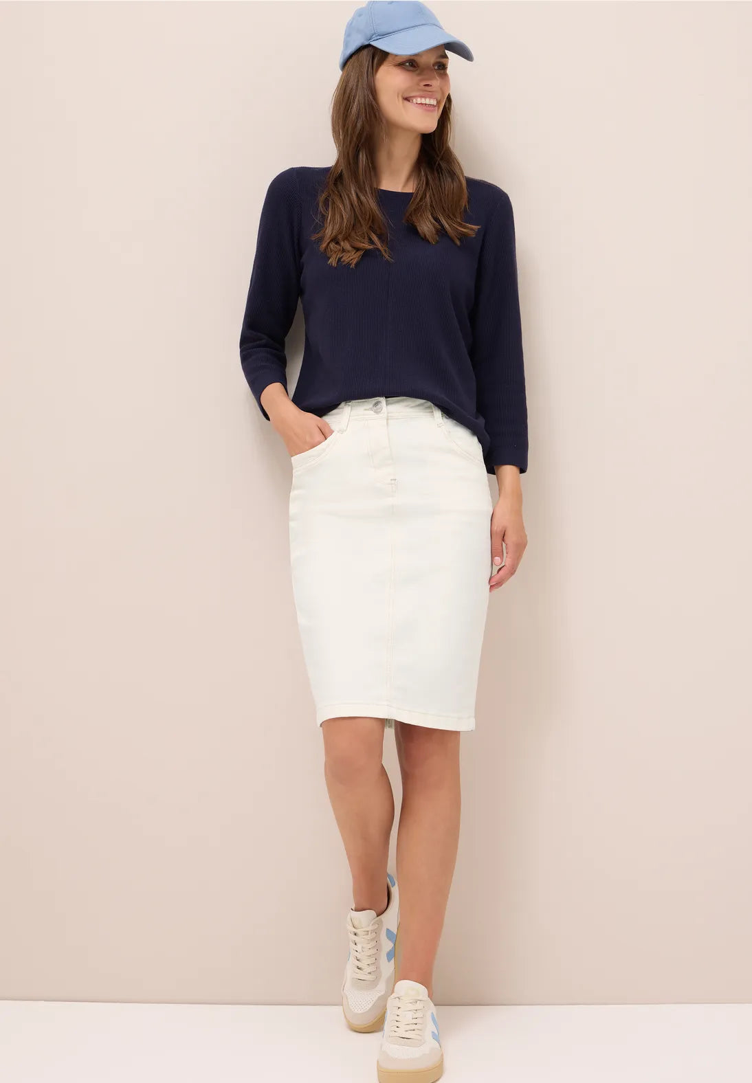 Woman wearing a navy sweater, white skirt, and blue cap against a beige background