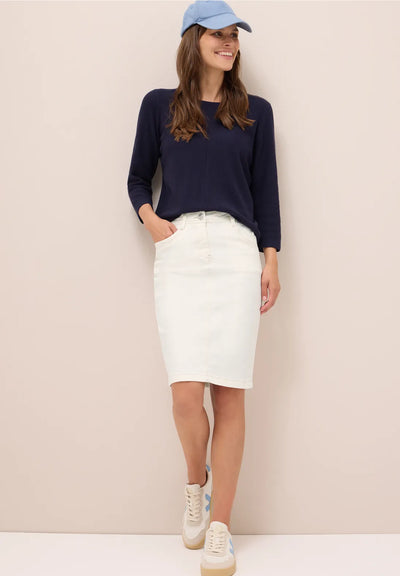 Woman wearing a navy sweater, white skirt, and blue cap against a beige background