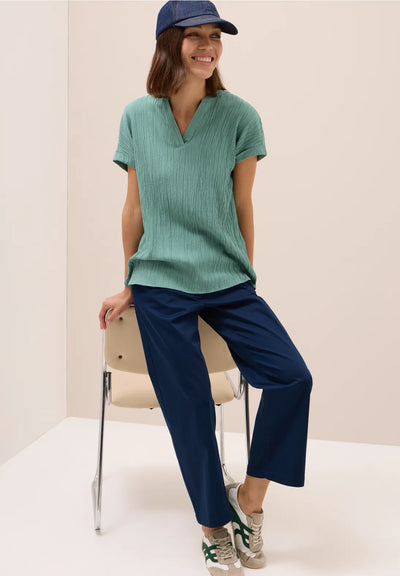 Woman wearing a green ribbed sweater and navy pants sitting on a chair against a plain background