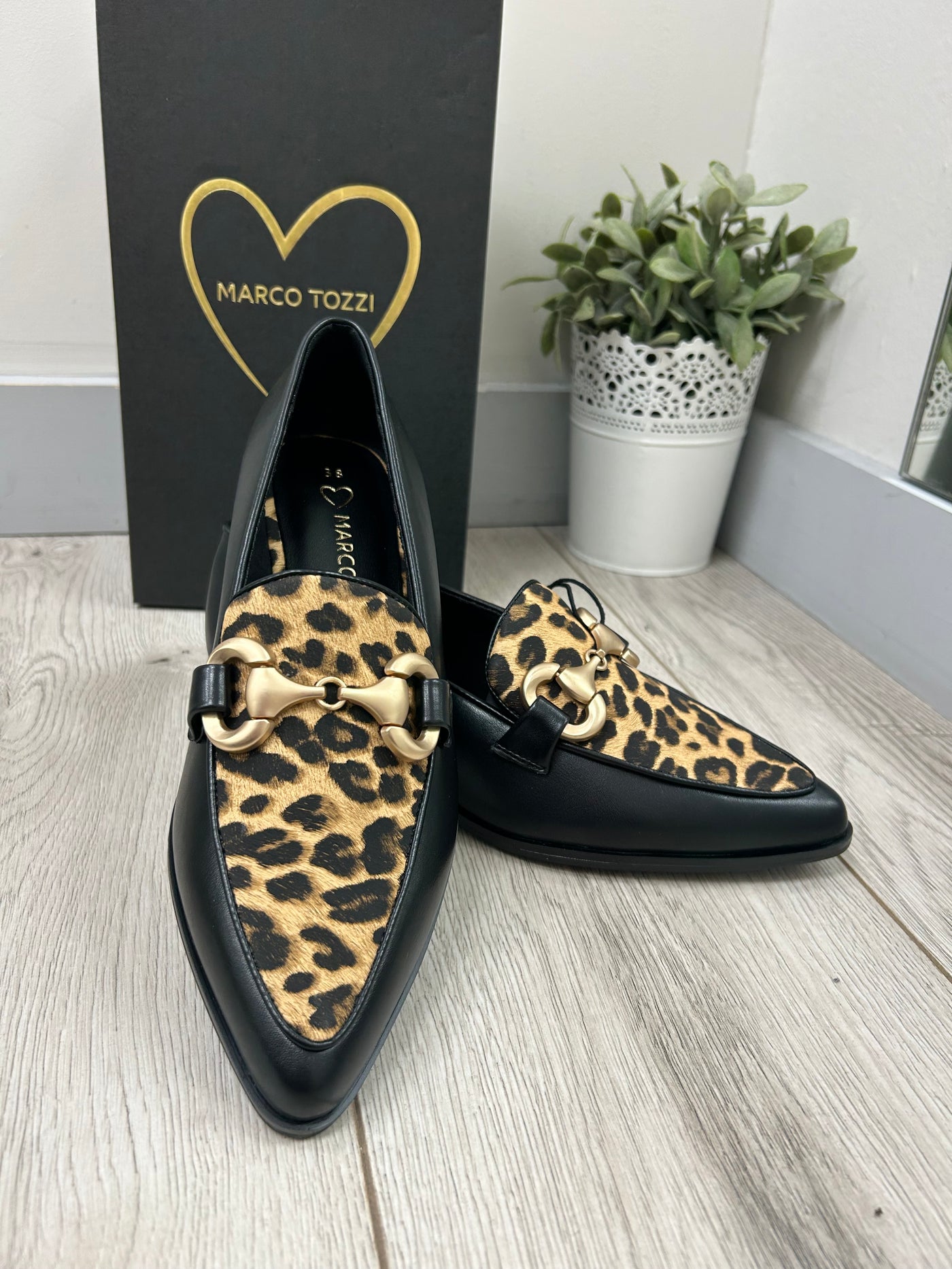 Black shoes with leopard print soles on a wooden floor next to a plant and Marco Tozzi box.