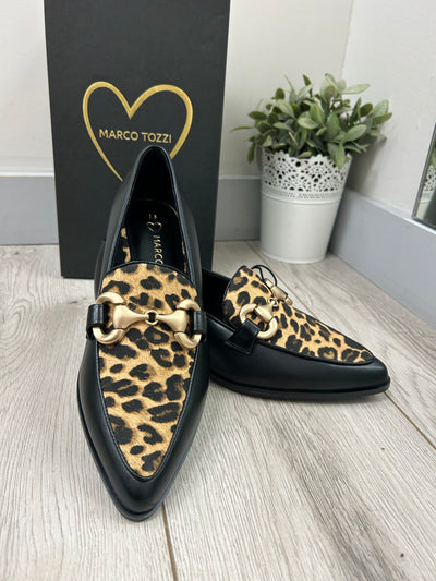 Black shoes with leopard print soles on a wooden floor next to a plant and Marco Tozzi box.