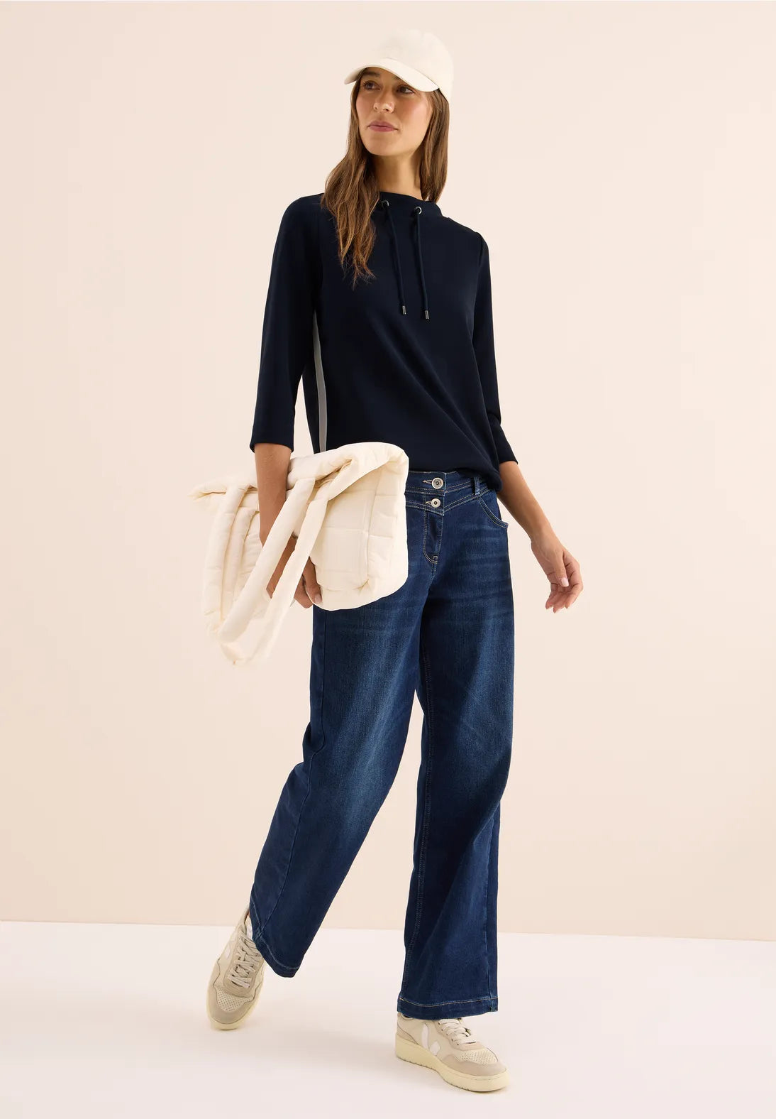 Woman wearing a navy sweater and blue jeans, holding a beige jacket against a light pink background