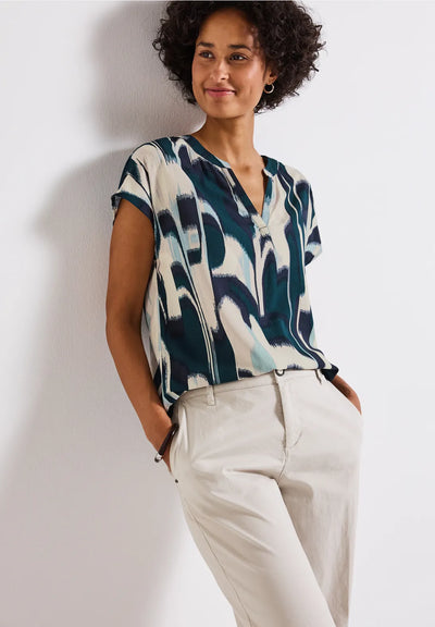 Woman wearing a patterned top and white pants against a plain background