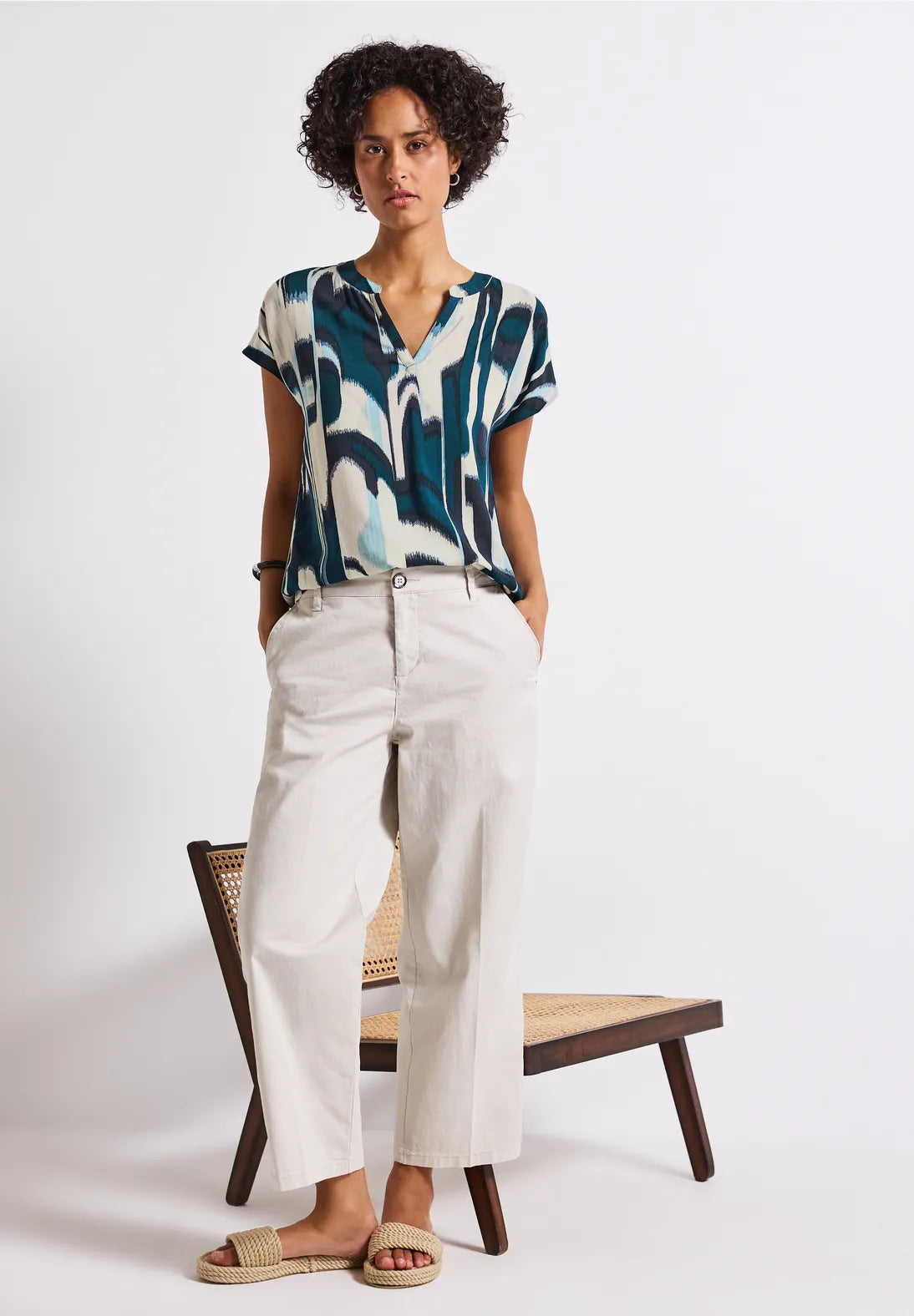 Woman wearing a patterned top and white pants standing in front of a white background
