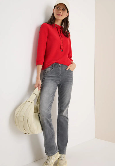 Woman wearing a red sweater and gray jeans, holding a beige bag against a white background