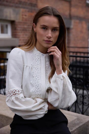 Woman wearing a white blouse with lace details against a brick wall.