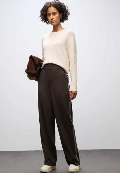 Woman wearing a white sweater and brown pants holding a brown jacket against a plain background