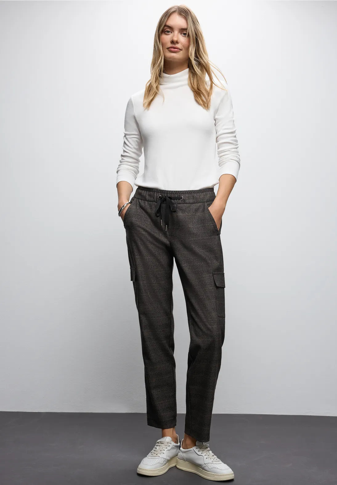 Woman wearing a white turtleneck and dark cargo pants against a plain background