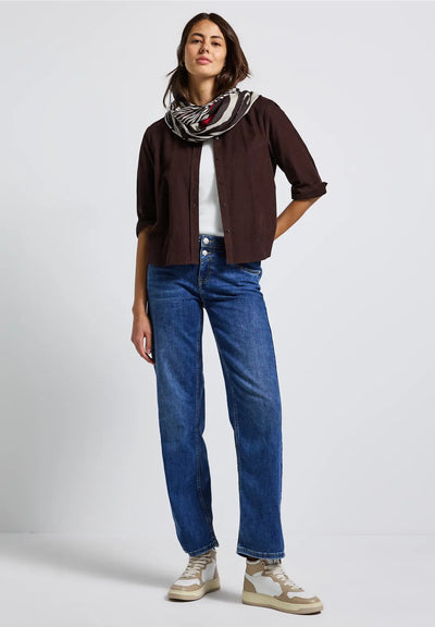 Woman wearing a brown shirt, white sneakers, blue jeans, and a patterned scarf on a light gray background