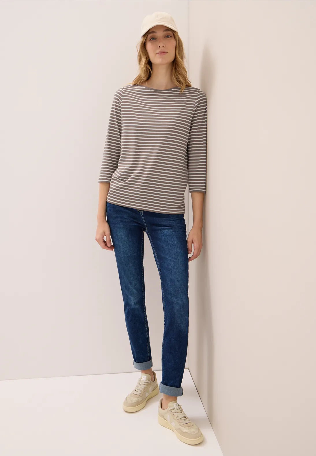 Woman wearing a striped sweater, blue jeans, and beige sneakers against a white wall.